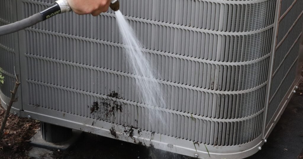 A Henry Heating and Cooling technician is cleaning an air conditioning unit with a hose.