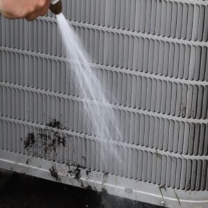 A Henry Heating and Cooling technician is cleaning an air conditioning unit with a hose.