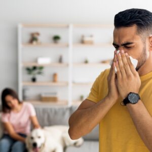 Man sneezing in a modern living room while a woman sits on a couch in the background with a dog.