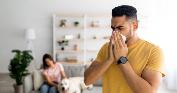 Man sneezing in a modern living room while a woman sits on a couch in the background with a dog.